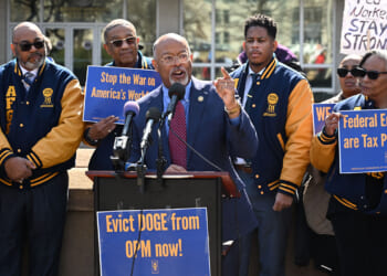 Protesters with signs from AFGE, the largest federal government union, stand together at a rally. A man speaks at a podium, surrounded by others holding signs with messages supporting workers and federal employees. Most are wearing matching blue and yellow jackets.