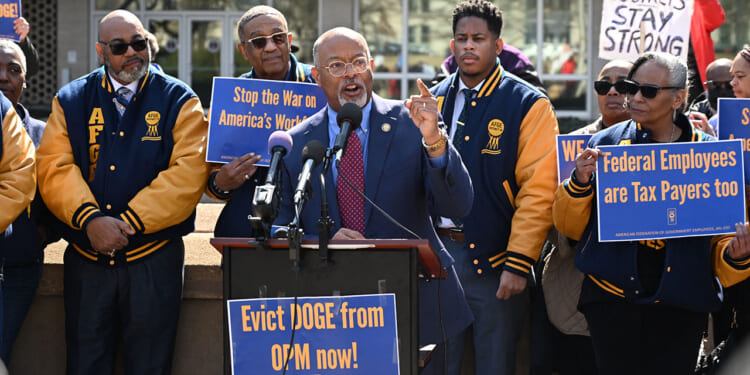 Protesters with signs from AFGE, the largest federal government union, stand together at a rally. A man speaks at a podium, surrounded by others holding signs with messages supporting workers and federal employees. Most are wearing matching blue and yellow jackets.