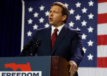 Ron DeSantis in a suit speaks at a podium labeled FREEDOM with a Florida outline, in front of a large American flag, emphasizing the importance of election integrity.