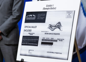 A sample ballot is displayed on an easel, labeled Exhibit 1, with a person in a suit standing nearby. The ballot includes official markings and return instructions.