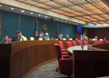 Members of the Joint Election Equipment Specifications and Standards Committee sit at a long, curved table at their third meeting with red chairs, U.S. and state flags, microphones, and nameplates.