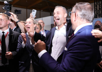 A group of people from the AfD in business attire cheer and celebrate together indoors. Some are smiling and shouting, while others take photos. The atmosphere is lively and joyful.