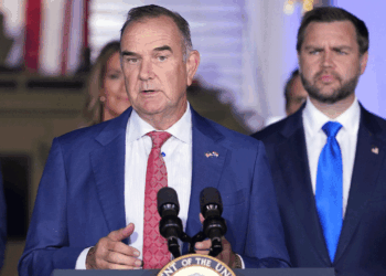 A man in a blue suit speaks at a podium with microphones, while another man in a suit stands behind him. An American flag is visible in the background, suggesting this event may be taking place in Missouri.