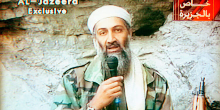 Osama Bin Laden in camouflage clothing and a white head covering speaks into a microphone against a rocky background, referencing the events of 9/11. Al-Jazeera Exclusive and Arabic text appear on the screen.
