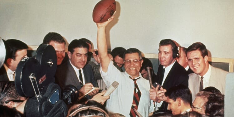 A smiling man in a tie triumphantly holds a football above his head, surrounded by reporters, cameras, and microphones in a crowded room.