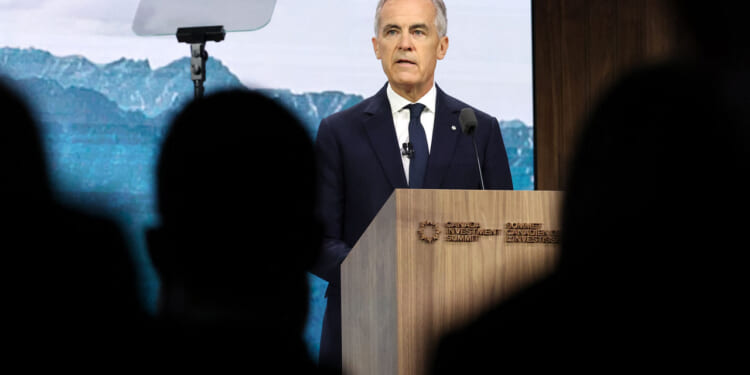 Mark Carney in a suit speaks at a podium during a summit in Canada, with a mountain backdrop and a teleprompter visible. Silhouetted audience members are in the foreground.
