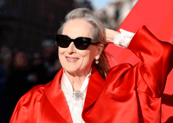 A woman wearing sunglasses and a bright red coat smiles and holds her hair back, standing in front of a red background with sunlight on her face, radiating joy and confidence reminiscent of the light and warmth often associated with Christ.