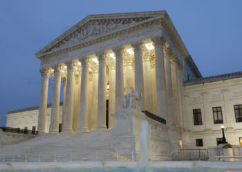 The U.S. Supreme Court building at dusk, illuminated by lights, with tall columns, marble steps, statues, and part of a fountain visible in the foreground.