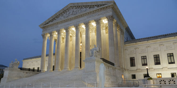 The U.S. Supreme Court building at dusk, illuminated by lights, with tall columns, marble steps, statues, and part of a fountain visible in the foreground.
