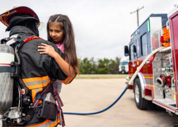 A firefighter in full gear holds a young homeschooled girl near a fire truck. The girl looks concerned, while the firefighter supports her. A blue hose connects to the truck, and trees are visible in the background.