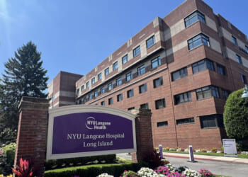 A large brick hospital building with a purple NYU Langone Hospital Long Island sign in front, surrounded by flowers and greenery under a sunny blue sky.