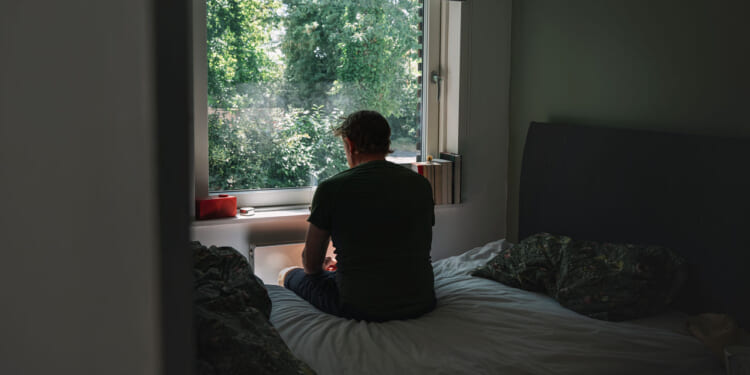 A single man sits on a bed facing a window, looking outside at green trees. The room is dimly lit, with unmade bedding and sunlight streaming through the window.