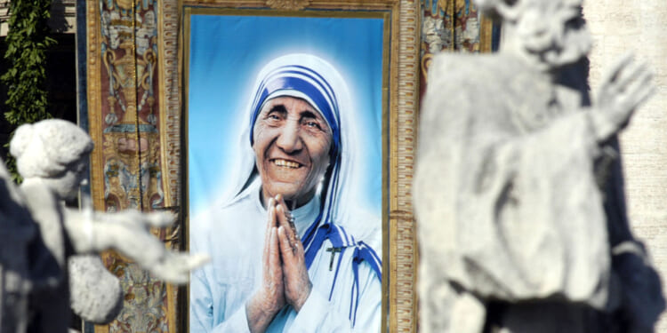 A large portrait of a smiling woman in a white and blue sari with hands clasped in prayer, framed and displayed outdoors, with blurred statues in the foreground.