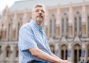 University of Washington professor Stuart Reges, who got disciplined over a land acknowledgment. Pictured: Reges, an older man in a blue plaid shirt sits outdoors, looking upwards thoughtfully. A large, historic building with arched windows and detailed stonework is in the blurred background.