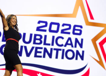 Monica De La Cruz in a black dress waves while standing in front of a large sign that says 2026 Republican Convention with a star graphic.