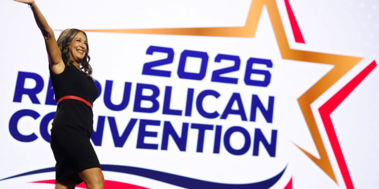 Monica De La Cruz in a black dress waves while standing in front of a large sign that says 2026 Republican Convention with a star graphic.