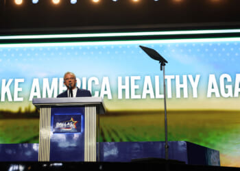 Robert F. Kennedy Jr. speaks at a podium on stage with a large screen behind them displaying the words MAKE AMERICA HEALTHY AGAIN. The podium has a Dallas sign on the front.