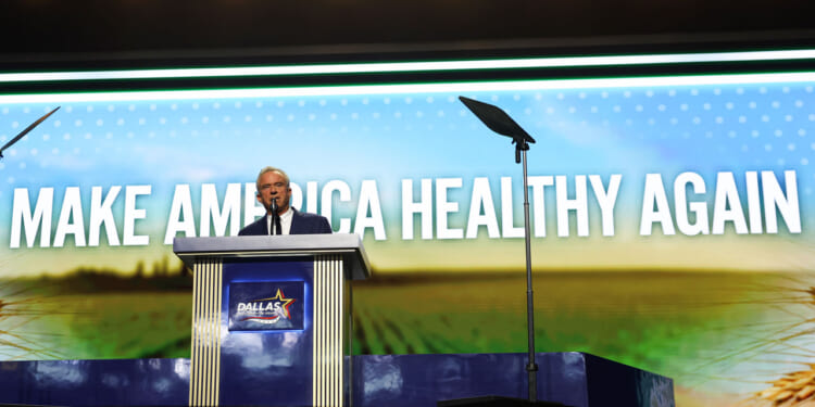 Robert F. Kennedy Jr. speaks at a podium on stage with a large screen behind them displaying the words MAKE AMERICA HEALTHY AGAIN. The podium has a Dallas sign on the front.