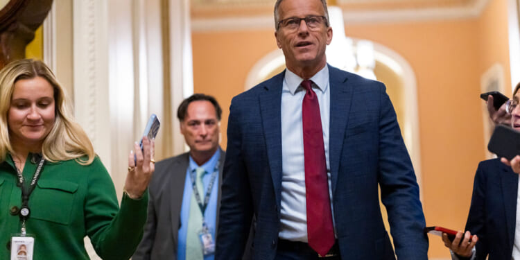 A man in a blue suit and red tie walks down a hallway with others; he is surrounded by people holding phones and wearing ID badges. The setting appears to be a government or official building.