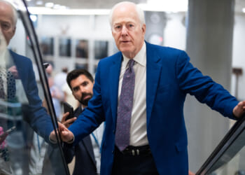 John Cornyn in a blue suit and patterned tie stands on an escalator, looking ahead with quiet resolve, while another man below—perhaps engaged in a discussion about socialism—holds a phone and looks up at him.