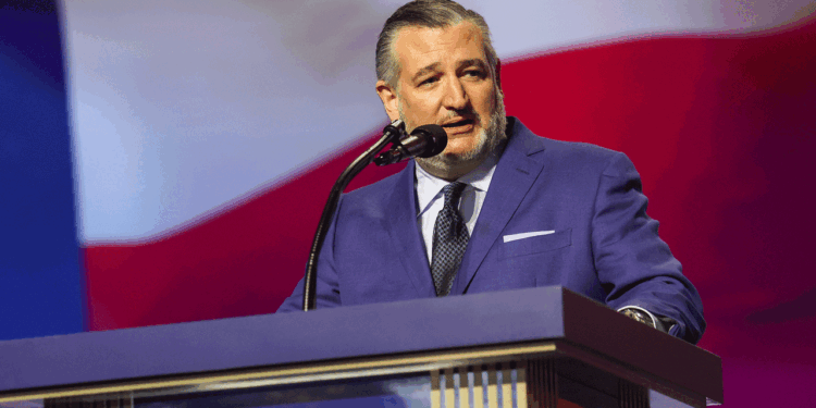 Sen. Ted Cruz in a blue suit speaks at a podium with a microphone, with a large red, white, and blue background behind him.