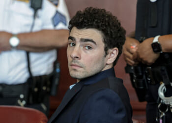 A man in a suit sits in a courtroom, looking over his shoulder. Two uniformed officers with handcuffs and radios stand nearby, slightly out of focus.
