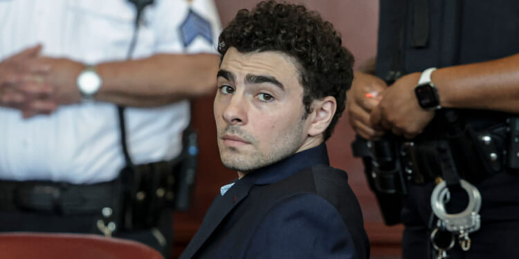 A man in a suit sits in a courtroom, looking over his shoulder. Two uniformed officers with handcuffs and radios stand nearby, slightly out of focus.