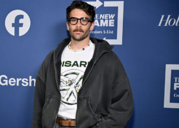 A man with dark hair, glasses, and a mustache stands in front of a blue backdrop with logos. He wears a white graphic t-shirt, a dark hoodie, and jeans.