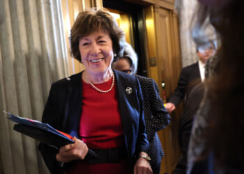 Sen. Susan Collins in a red dress and black blazer, smiling, holds folders while standing near an elevator in a building with marble columns. Other people are around her.
