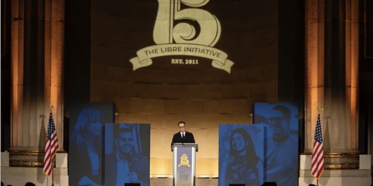 A speaker stands at a podium with The Libre Initiative Est. 2011 projected above. Blue portraits and two American flags flank the stage in a grand hall.