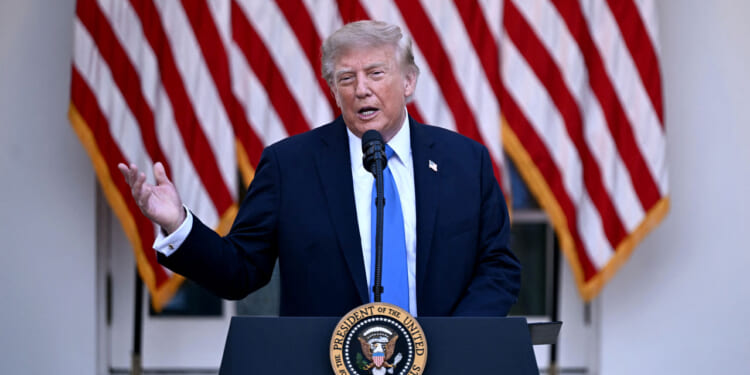 President Donald Trump in a suit speaks at a podium with the U.S. presidential seal, in front of several large American flags.