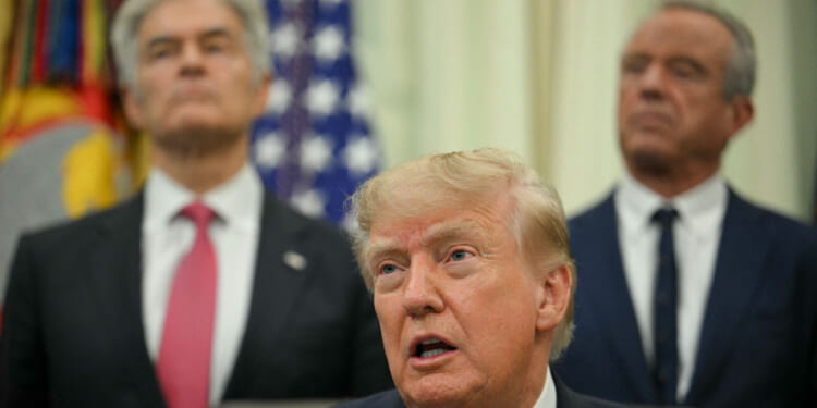 Three men in suits stand indoors; the man in front is speaking, while two others stand behind him. U.S. flags and official emblems are visible in the background.