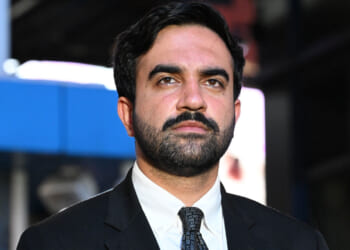 A man with dark hair and a beard wears a black suit, white shirt, and patterned tie, standing outdoors with a blurred city background.