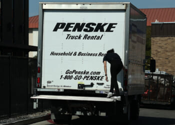 A person climbs onto the back of a white Penske moving truck parked near a building during daylight. The truck advertises household and business rentals, with contact information and a website visible, suggesting it’s ready to support any move—whether for an individual or a union organizing collective effort.