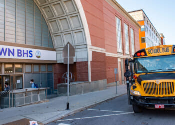 A yellow school bus is parked outside a brick school building labeled DOWNTOWN BHS, while students enter through the main doors. The weather is clear and sunny.