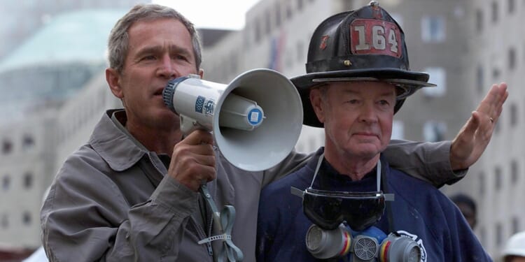 A man in a jacket speaks through a megaphone while standing with his arm around a firefighter wearing a helmet marked 164 and a respirator around his neck. Buildings are visible in the background.