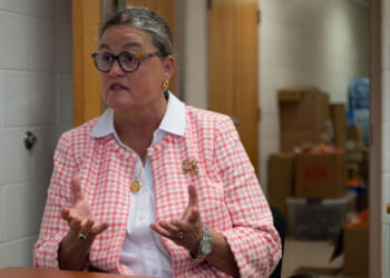 Michelle Reid with glasses and gray hair sits in an office, wearing a pink and white checkered jacket, gesturing with her hands while speaking. Boxes and office supplies are visible in the background.