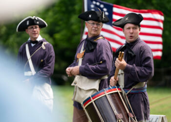 Three men in historical military uniforms stand outdoors with an American flag behind them. One plays a drum, while the others watch. The scene appears to be a reenactment.