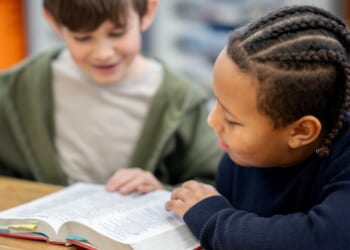 Two children sit at a classroom table sharing and reading a Bible.