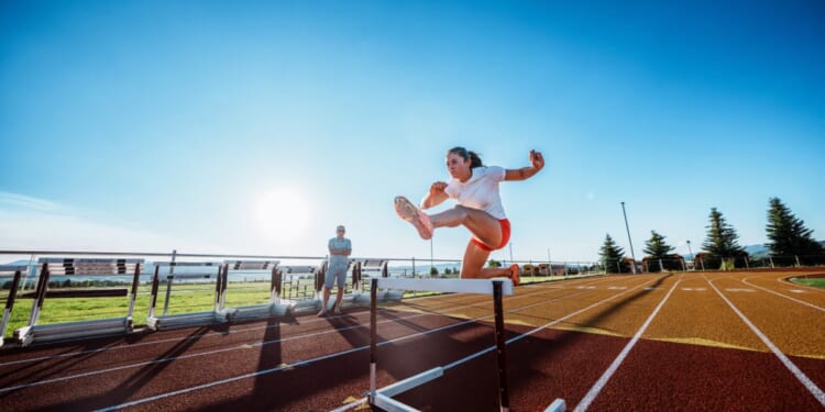 A female collegiate athlete powers over a hurdle at full speed during an intense solo training session with her coach observing nearby.